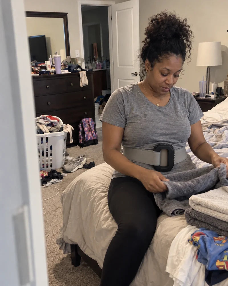 A mom folding laundry on the edge of her bed in a messy bedroom while wearing the Restora Core Wrap over her T-shirt