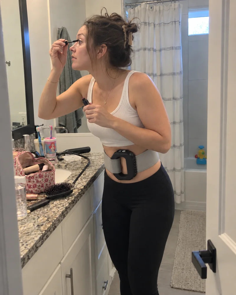 A mom doing her makeup in the bathroom mirror while wearing the Restora Core Wrap at her waist